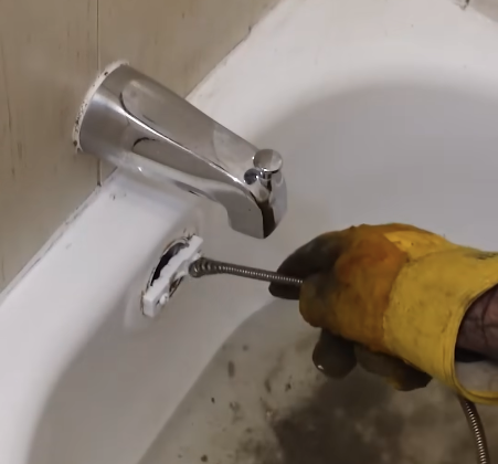 Gloved hand running a drain snake cable through a bathtub overflow to clear a slow tub drain