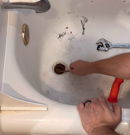 Plumber clearing a clogged bathtub drain with the strainer removed and tools set on the tub