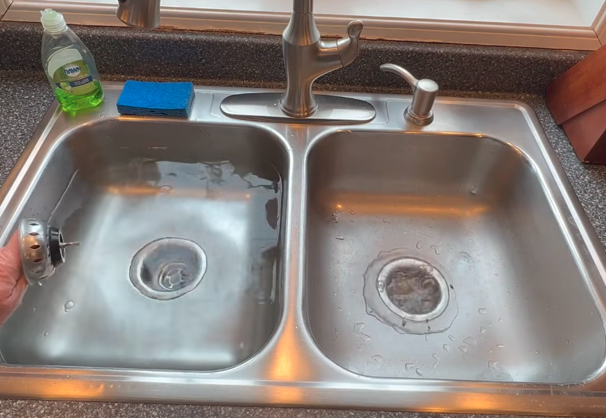 Double kitchen sink with standing water in one bowl and the basket strainer pulled, a clogged kitchen sink drain