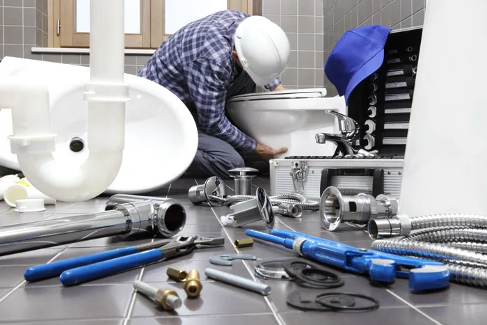 Plumber in a hard hat working on a toilet with pipes and wrenches nearby.